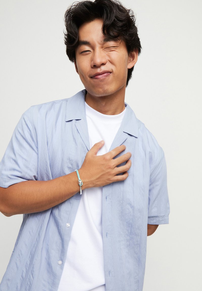 Chemise à manches courtes bleu clair, à boutons et avec une coupe décontractée, portée sur un T-shirt blanc. Tissu texturé remarquable et design simple accentué par un bracelet.