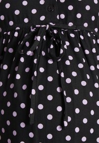 Vestido de tecido preto com bolinhas brancas, apresentando uma cintura franzida com um detalhe de laço e fecho de botão. Design texturizado e casual.