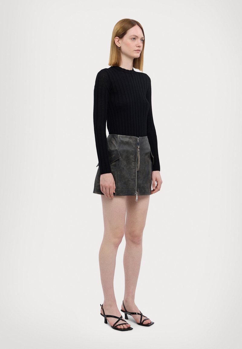 Maglia nera a coste a maniche lunghe abbinata a una gonna in pelle grigia con zip e due tasche laterali. La modella indossa tacchi neri con cinturini.
