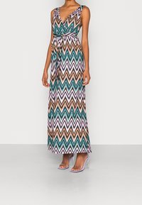 Robe maxi avec un motif en chevron turquoise, orange et rose. Décolleté en V profond, sans manches, taille nouée, accompagnée de talons transparents.
