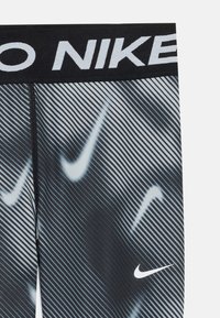 Kadar na crno-sive Nike tajice s dijagonalnim prugama i ponovljenim bijelim Nike swoosh logotipima, uz crni pojas s natpisom "NIKE".
