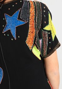 Svart tyg med färgglada paljetter i stjärn- och kometmönster i blått, gult, orange och grönt på ett plagg med korta ärmar.