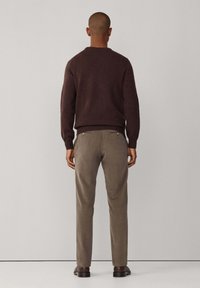 Mann, der einen dunkelbraunen Pullover, taupefarbene Hose und dunkelbraune Schuhe trägt, steht vor einer schlichten hellgrauen Wand.
