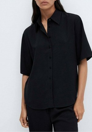 Femme portant une chemise noire ample à manches courtes avec col pointu et un pantalon noir assorti, sur fond blanc.