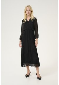 Schwarzes, plissiertes Maxikleid mit transparenten langen Ärmeln, einem Wickelausschnitt und einer taillierten Taille. Kombiniert mit schwarzen Slingback-Schuhen mit spitzer Zehenform.