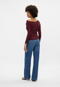 Bordo garas piedurkņu blūze ar izgrieztu aizmuguri, kombinēta ar zilām platām biksēm. Audumam ir gluda tekstūra un pieguļoša forma.