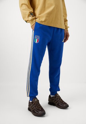Blauwe sweatpants met gouden zijstrepen, voorzien van een geborduurd Italië-vlag embleem. Gecoördineerd met bruine sportschoenen.