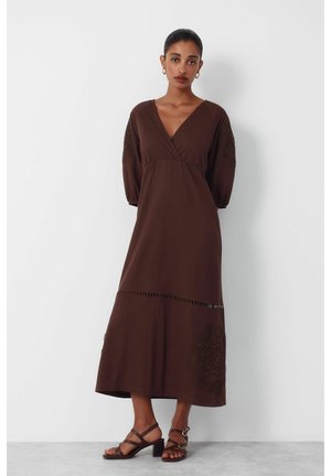 Frau trägt ein braunes Midi-Kleid mit 3/4-Puffärmeln und Spitzendetails an Ärmeln und Rock, kombiniert mit braunen Riemchen-Sandaletten mit Blockabsatz.