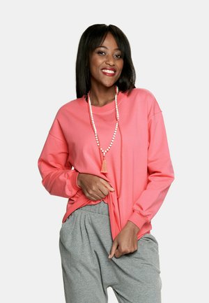 Korali sweatshirt z długim rękawem, luźny krój, zestawiony z szarymi dresami. Naszyjnik z koralików dodaje detali do stroju.