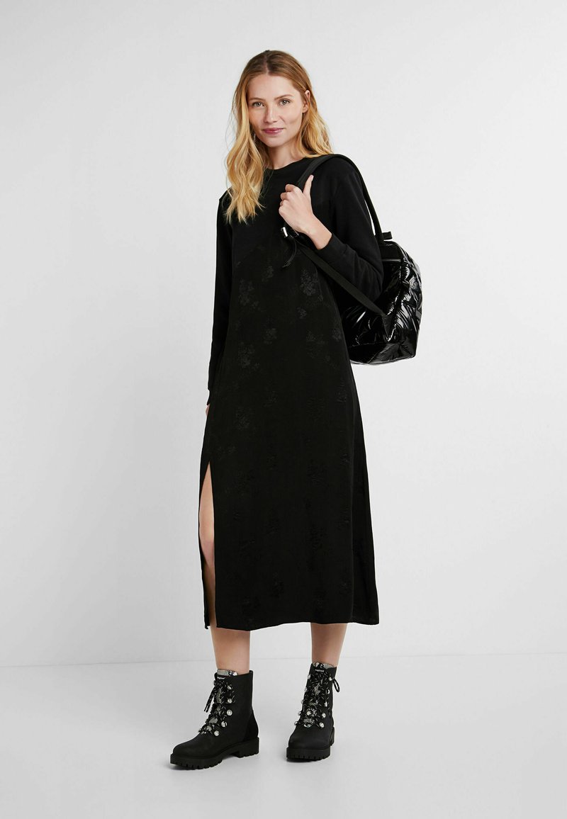 Zwarte langemouwen jurk met een bloemenpatroon, een split aan de zijkant, gecombineerd met zwarte veterschoenen en een glanzende zwarte rugzak. Effen achtergrond.