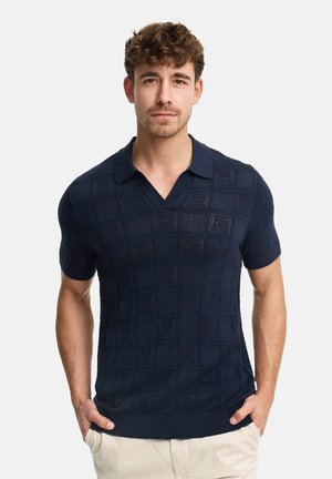 Hombre con polo de manga corta azul marino de punto con patrones geométricos, de pie con las manos en los bolsillos del pantalón beige, fondo neutro.