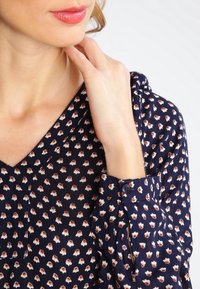 Marineblouse met een wit en roestkleurig bloemenpatroon, V-hals, met knopen en van lichte stof. Close-up van een hand op de halslijn.