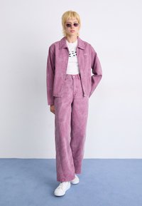 Rosa denimjakke og bukser med slitt utseende, kombinert med en hvit t-skjorte med grafikk og hvite sneakers. Runde solbriller fullfører antrekket.