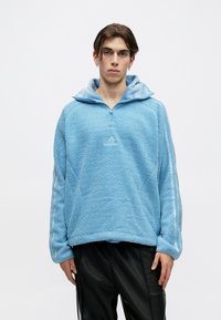Giovane con capelli corti castani, che indossa un pullover in pile azzurro chiaro con mezza zip e pantaloni neri, in piedi su uno sfondo bianco.