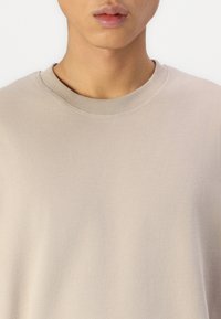 Béžová mikina s kulatým výstřihem, jemnou texturou a ribbovanými lemy. Detailní pohled zdůrazňuje minimalistický design a hladký povrch tkaniny.