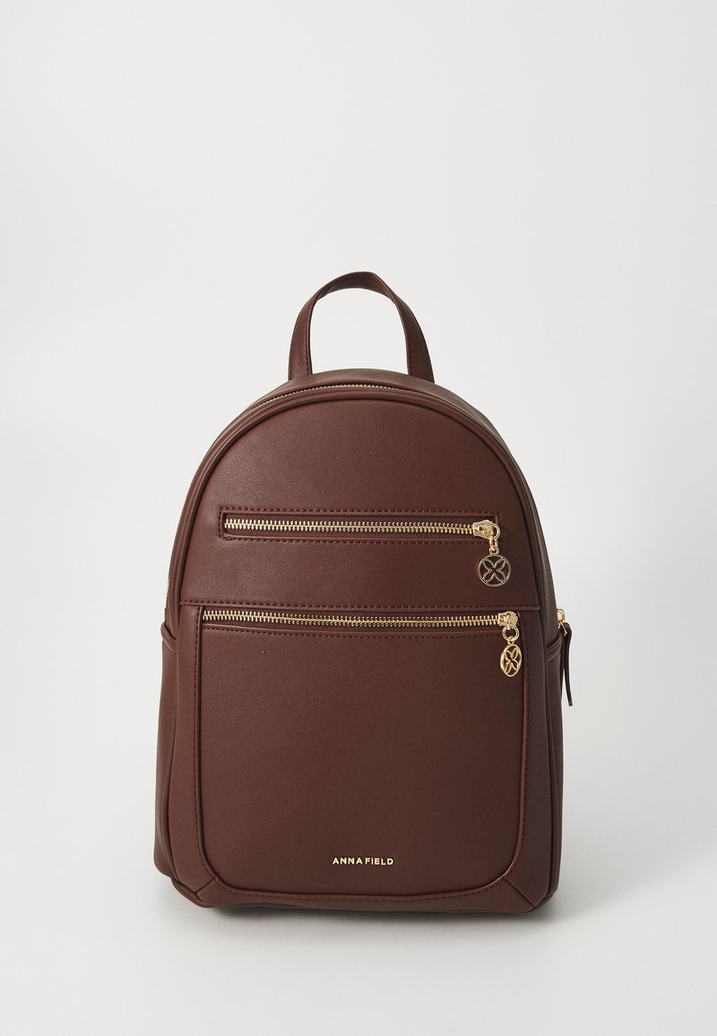 Sac à dos en simili cuir marron avec une forme arrondie, doté de deux fermetures éclair à l'avant, d'accents dorés et d'une poignée supérieure pour le transport.