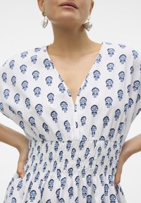 Weißes Kleid mit blauen Blumenmustern, V-Ausschnitt, kurzen Ärmeln und geraffter Taille. Es zeigt lange Ohrringe und hat eine glatte Stofftextur.