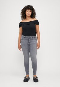 Fekete, vállra lógszó bodyscrap, drapált dekoltázzsal, szűk szürke farmerral párosítva. A modellen fekete platform cipők vannak, és egy világos háttér előtt áll.