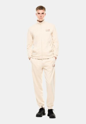 Giovane uomo in piedi che indossa una tuta beige EA7 con top con zip e pantaloni abbinati, abbinata a scarpe da ginnastica nere, mani nelle tasche.