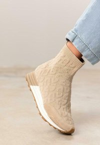 Zapatilla deportiva de estilo calcetín en color beige con parte superior de punto texturizado, entresuela blanca y suela de goma marrón, que presenta un patrón de logo distintivo.