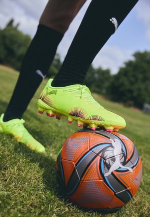 Svetlo zeleni nogometni čevlji s teksturiranimi stranicami in rožnatimi nogicami, postavljeni nad vzorčasto oranžno in črno nogometno žogo na travi.
