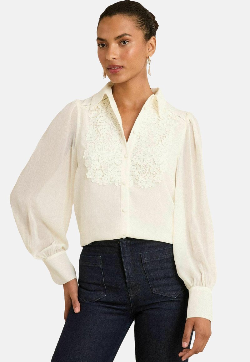 Blusa bianca con maniche lunghe a sbuffo e un dettaglio in pizzo sul davanti. Con bottoni, colletto e polsini, indossata con jeans scuri.