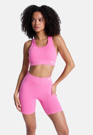 Femme aux cheveux bouclés portant un soutien-gorge de sport côtelé rose vif assorti à un short de cyclisme, posant avec une main sur la hanche sur un fond blanc.