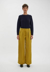 Pull en maille bleu marine à manches longues associé à un pantalon large taille haute en moutarde ; mannequin portant des chaussures noires ; fond uni.