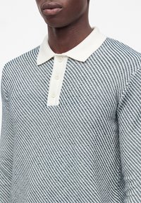 Homme portant un polo en maille à manches longues avec un col blanc et un tissu à motif diagonal bleu et blanc.