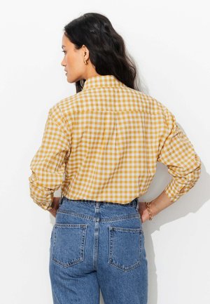 Femme aux longs cheveux foncés portant une chemise vichy jaune et blanche rentrée dans un jean bleu taille haute, debout mains sur les hanches.
