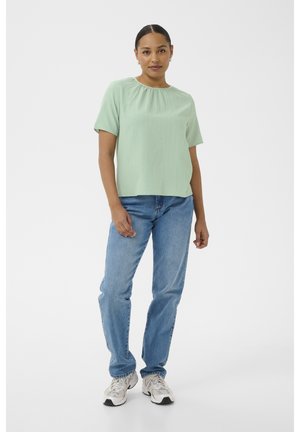 Žena oblečená v zelenkavě světle krátkém rukávu, modrých džínách a bílých teniskách, stojící na jednoduchém bílém pozadí.