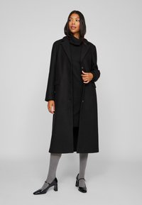 Pikk must villane mantel, millel on lõigatud krae, ühe nuppe sulgemine ja ees taskud, kantud musta kõrge kaelusega kleidi ja hallide sukkpükstega.