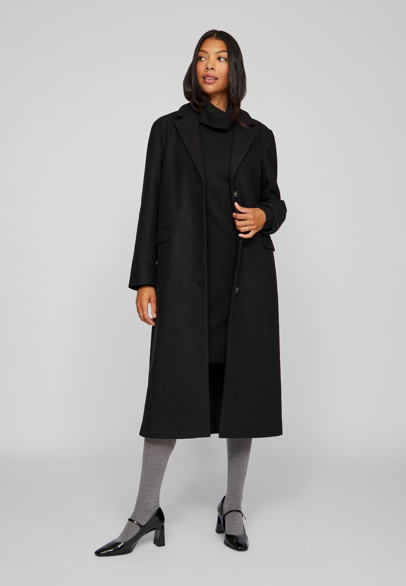 Pikk must villane mantel, millel on lõigatud krae, ühe nuppe sulgemine ja ees taskud, kantud musta kõrge kaelusega kleidi ja hallide sukkpükstega.