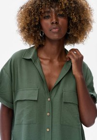 Vrouw met krullend haar die een groene dichtgeknoopte blouse draagt en de kraag vasthoudt, tegen een effen witte achtergrond.