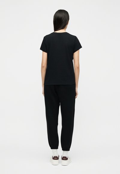 Γυναίκα όρθια με πλάτη, φοράει μαύρο κοντομάνικο μπλουζάκι, μαύρο παντελόνι jogger, λευκές κάλτσες και λευκά αθλητικά παπούτσια με μπορντό τακούνια.