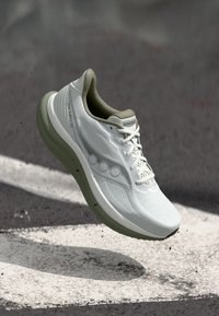 Λευκό παπούτσι τρεξίματος Saucony Triumph 20 στον αέρα πάνω από άσφαλτο με λευκή βαμμένη γραμμή, που δείχνει πλευρικές και κορυφαίες λεπτομέρειες.