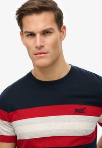Young man with short brown hair wearing a navy, red, and white striped t-shirt with a small embroidered logo on the chest.