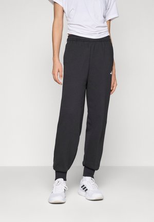 Person iført sorte joggebukser, hvide sneakers med sorte striber og en indstøbt hvid t-shirt, der står mod en ensartet lys baggrund.
