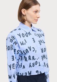 Světle modrá croptop košile s límečkem a výrazným černým textem různých velikostí po celé látce. Hladký povrch, zapínání na knoflíky.