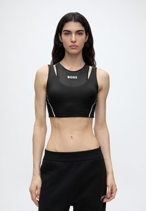 Kvinde med langt mørkt hår iført en sort sport crop top med "BOSS" logo og sorte joggingbukser mod en ensfarvet hvid baggrund.