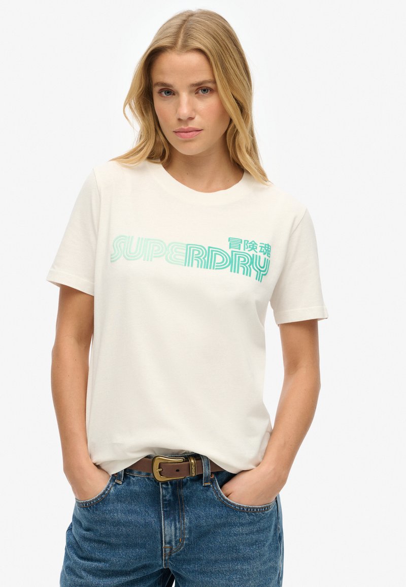Kremowa koszulka z bawełny z turkusowym napisem "SUPERDRY". Klasyczny okrągły dekolt i krótkie rękawy. Nosi ją z niebieskimi dżinsami.