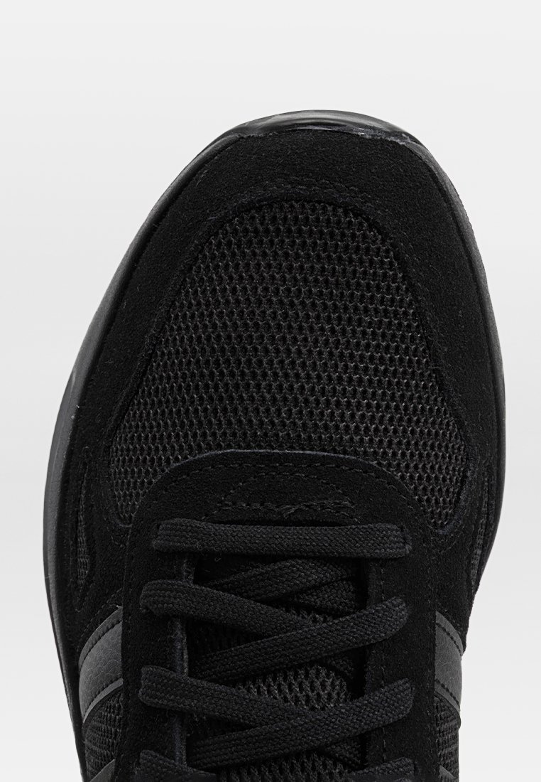 Scarpa sportiva nera con tomaia in rete testurizzata, dettagli in suede e lacci neri piatti. Include sottili strisce grigie sul lato.