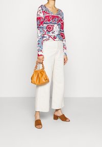Haut à manches longues et col en V avec des motifs floraux rouges, bleus et blancs, associé à un pantalon large beige clair, des mules à talons carrés en daim marron et un sac à main orange.