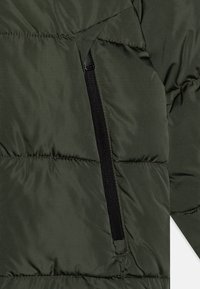 Grön puffjacka med quiltad textur, med en svart dragkedjeficka på vänster sida, tillverkad av lätt syntetiskt material.