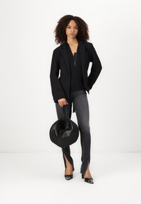 Črn blazer preko črnega topa, v kombinaciji s sivo jeans tesnilo z razporkom. Drži okroglo črno torbico in nosi črne elegantne čevlje.