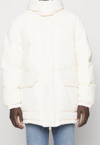 Bílý zimní kabát s vysokým límcem, předními kloboukovými kapsami a elastickými manžetami, nošený přes modré džíny.