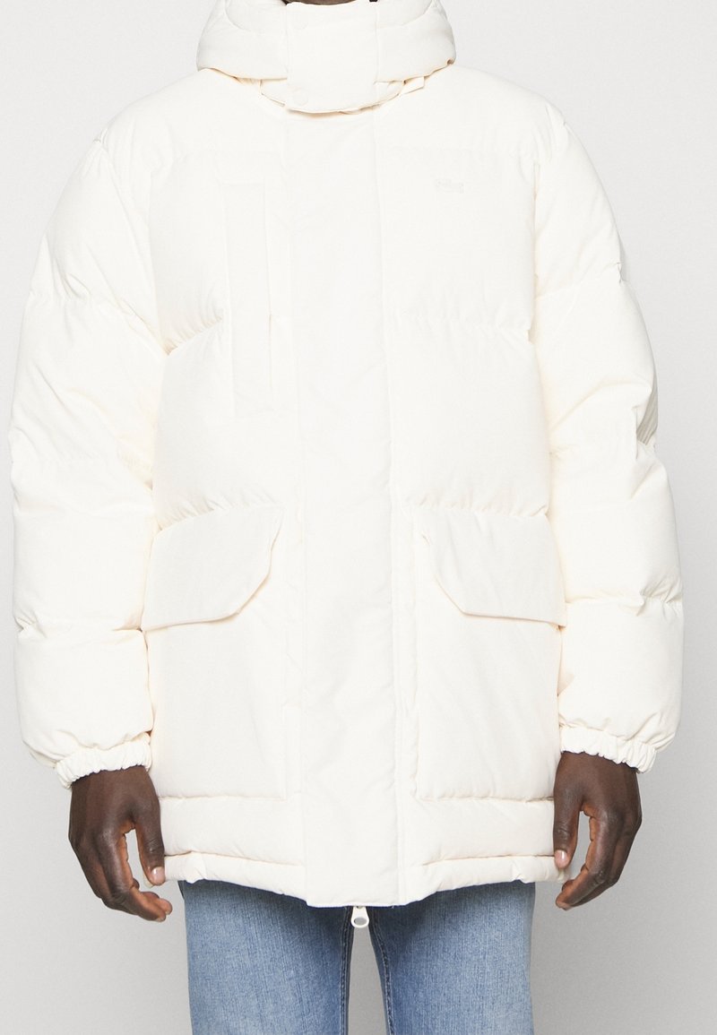 Bílý zimní kabát s vysokým límcem, předními kloboukovými kapsami a elastickými manžetami, nošený přes modré džíny.