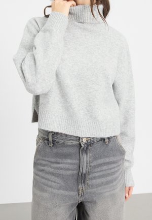 Personne portant un pull à col roulé côtelé gris clair et un jean gris délavé taille haute, la main touchant le cou.
