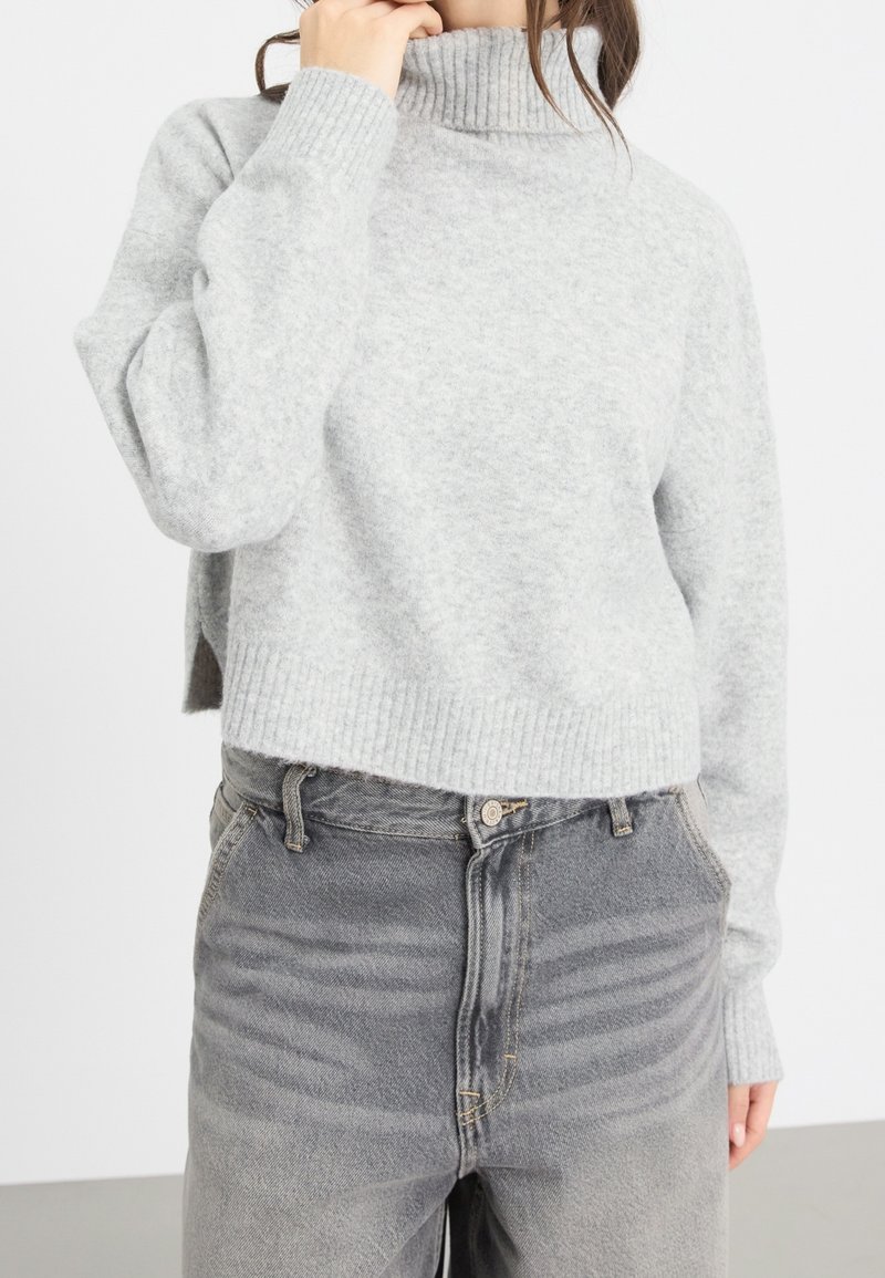 Personne portant un pull à col roulé côtelé gris clair et un jean gris délavé taille haute, la main touchant le cou.