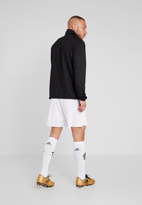 Svart langermet treningsjakke, hvite shorts og gull soccer sko med svarte detaljer; modellen står med ryggen til betrakteren, sokker til midten av leggen.